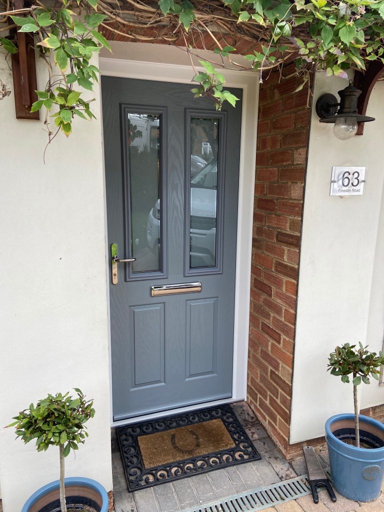 A Silver Grey Composite Door set in to a red brick wall with a horseshoe doormat and two pot plants outside.